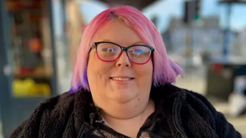 Ms Owen is smiling at the camera while wearing glasses that have a black frame around the top and a pink frame on the bottom, with a clear lens in the centre. She has medium length pink hair that is purple on the ends. She is wearing a black jacket and the background has is out of focus.