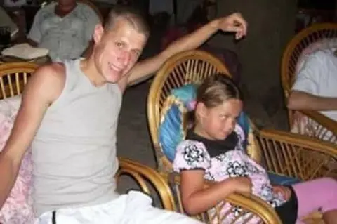 Courtney Vernon A young man wearing a grey vest and white shorts is sitting in a wicker chair and leaning towards his young daughter, who is wearing a flowery pink dress and sitting in a similar chair. Both are smiling.