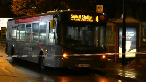 A bus next to a bus stop in the dark, with its lights on and blue and white police tape in front of it