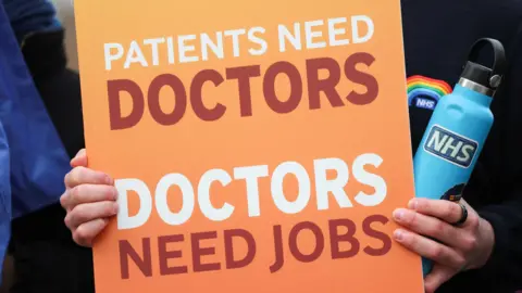 Reuters A doctor on a picket line holds an NHS branded water bottle and a sign which reads "Patients need doctors, doctors need jobs."
