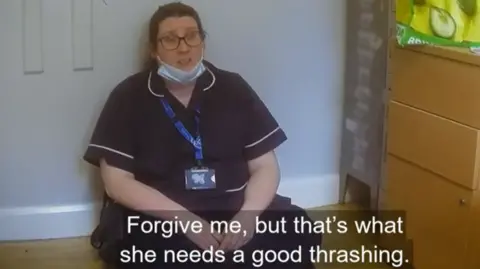 BBC Panorama A nurse, with dark blonde hair tied back, sits on a wooden floor inside a room. She is wearing a dark blue uniform, has a lanyard around her neck and has a face mask below her chin. 