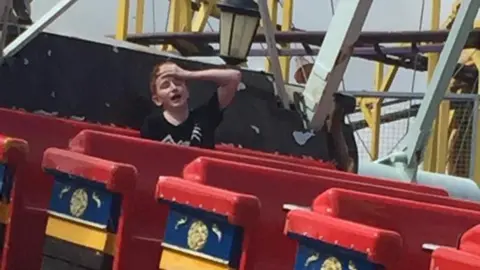 A boy holds his head and has his eyes closed as he sits on a pirate ship ride at a fairground 