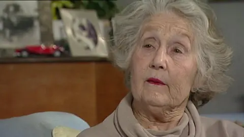 An elderly Noreen Riols gives a TV interview in a living room. She wears make-up and earrings, and her wavy hair is light grey.