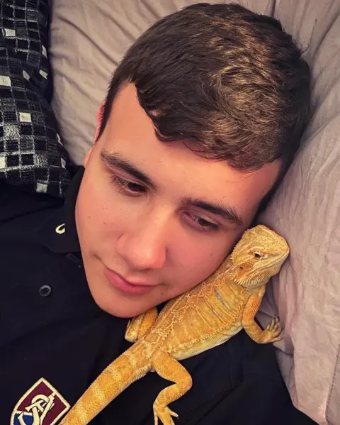 Naomi Rees-Issitt Teenage Jamie lying against some cushions with a bearded dragon propped up next to his face. He is wearing a dark top with a dark red, gold and blue logo on it.