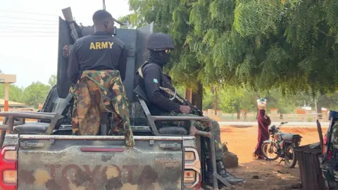 An army patrol truck, with two soldiers on the back carrying guns. To the right of the vehicle, a woman balancing a bucket on her head walks past a motorcycle