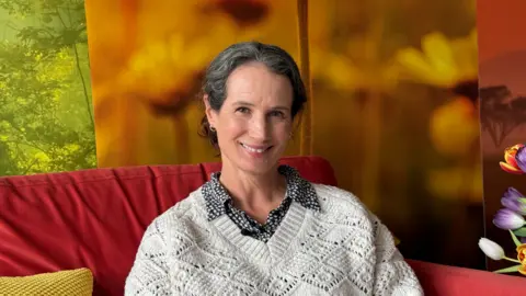 A woman sat on a red sofa. She is wearing a cream jumper and a patterned shirt underneath. She has greying dark short hair and is smiling at the camera.