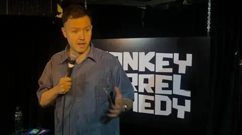 A comedian stands on a darkened stage with obscured white lettering on the back wall.