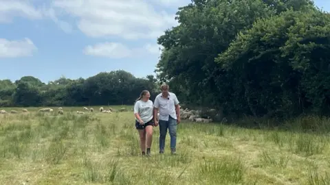 The image shows a man and a woman (Michael Jefferies and Annie Taylor) holding hands as they walk through a grassy field. Sheep are grazing behind them.
