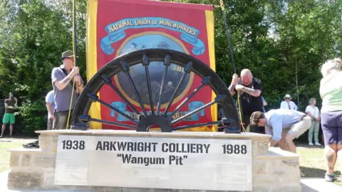 Two men hold a red miners banner behind a large half-wheel set in a stone base 