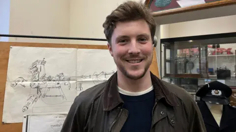 Joseph Rose - a young man with short, thick brown hair and a neatly trimmed beard - smiles as he stands in a museum surrounded by exhibits. He is wearing a dark wax jacket and a blue jumper. Behind him, a diagram of a machine is pinned to a board. To the right, a military uniform stands in front of a display case.