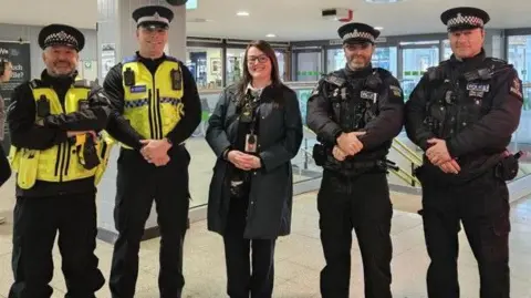 Four police officers in uniform - two from Devon and Cornwall Police and two from British Transport Police - stand either side of a woman inside a train station