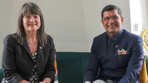 Cranfield University A woman and a man sitting on a blue sofa who are both smiling looking at the camera. The woman (left) is wearing a brown jacket and a black and white dress. She has long, brown hair. The man (right) is wearing a blue jacket and shirt. He has glasses and short, black hair.