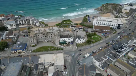 Aerial view of a coastal town featuring a mix of residential houses, commercial buildings, and a large hotel or apartment complex near the beach. Winding roads with parked cars run through the town. The ocean is in the background with waves crashing onto the shore.