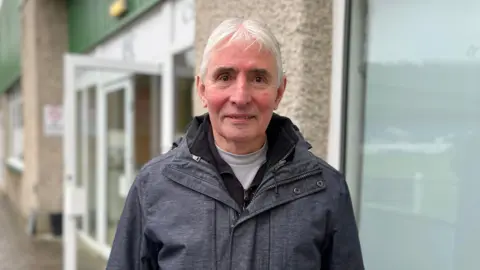 A grey-haired man with a grey jacket looks at the camera. Behind him are doors to a function suite