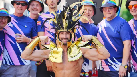 PA A person at the front is showing off their muscles and smiling at the camera whilst wearing a hold and black head piece and shoulder piece. They areas surrounded by people wearing pink and blue tops.