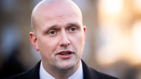 Stephen Flynn has a clean shaven head and is standing outdoors. He is wearing a black coat over a white shirt.