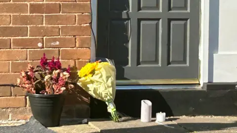 A bouquet of yellow flowers and two plastic candles sit on the doorstep of a house, next to a plant pot