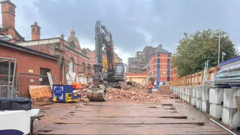BBC A fenced off building site where a large digger is parked on a pile of bricks and rubble. Leicester's Victorian rail station can be seen in the back ground.