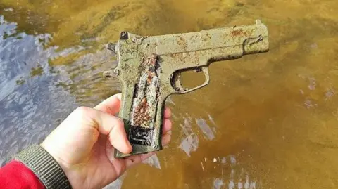 Calum Maclean A hand holding a rusty grey air pistol with murky river water in the background. 
