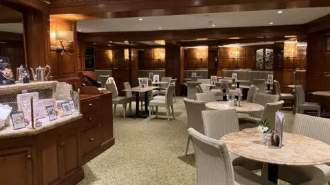 Kit Taylor/BBC A cafe with tables and chairs, dark wooden walls, and some signs and menus showing what food can be ordered. 