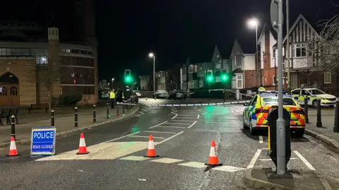 Orange traffic cones in a row on the street with a blue Police Road Closed sign. There are two police cars parked up at the side of the road and a square police cordon has been installed.