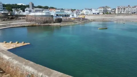 A picture of Havre des Pas bathing pool at high tide. The Lido buildings are visible to the left and there is a promenade of houses in the far distance. 