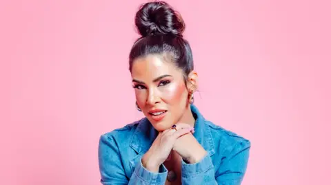 Aimie Atkinson Aimie Atkinson wearing a blue denim jacket with her dark hair in a bun is sat resting her face on her hands against a pink backdrop