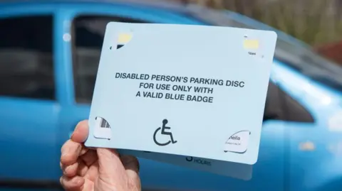 A woman's hand holding a Blue Badge parking disc issued in the UK to disabled drivers with a light blue car behind it. 