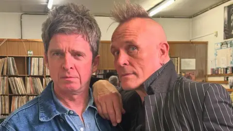 Noel Gallagher is standing next to John Robb in a vinyl shop. John is leaning on Noel's shoulder and both men are staring directly at the camera. Liam is wearing a denim jacket and denim shirt while John is wearing a pinstripe suit and shirt with the collar up. 