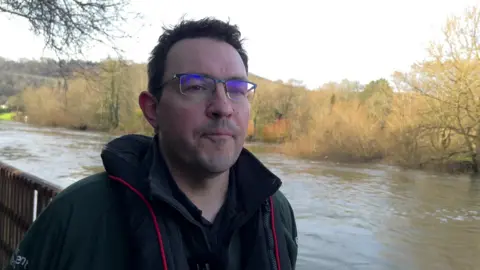 Stuart Manwaring has short black hair and glasses. He is wearing a black coat over a black fleece. Behind him is the river Thames.