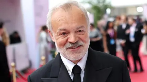Graham Norton at the Baftas 2026 on the red carpet, wearing a black suit and tie and a white shirt with diamante detail on the collar