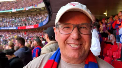 A man in glasses and a Crystal Palace scarf and hat smiles at the camera.