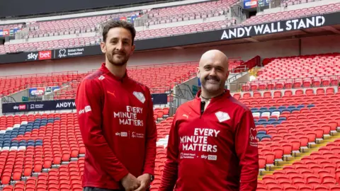 The Sky Bet & BHF Every Minute Matters Campaign Tom Lockyer and Andy Wall standing in front of the Andy Wall Stand inside Wembley Stadium.
