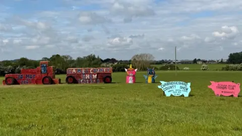 Soham YFC A tractor pulling a trailer, both made out of straw bales and painted red sitting in a grassy field. Behind it are two animal sculptures, one bright pink and the other blue and yellow. In front of that are two pig-shaped boards, one saying Cambridgeshire County Show and the other saying Soham Young Farmers. 