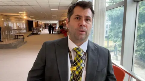 Lee Trewhela / LDRS Leigh Frost, a middle-aged man with short dark hair and light stubble, in a suit with a yellow, black and blue Cornish tartan tie.