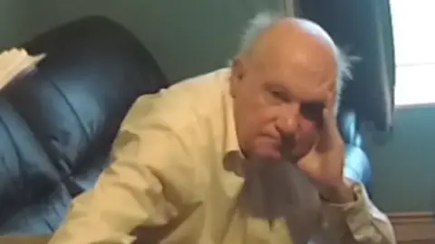 Raymond Rackley, sat on a brown leather sofa, holding his head on his left hand, is mostly bald with white hair around his head.