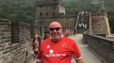 A man dressed in a red football shirt wearing sunglasses, standing in front of tourists on the Great Wall of China