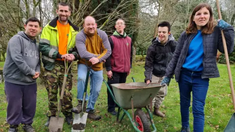 Five men and a woman standing in a garden holding gardening tools. There is a wheelbarrow next to them