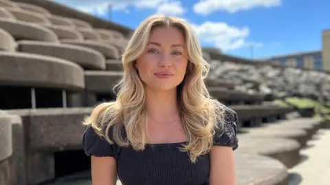 Daisy is sitting on curved concrete seating steps outdoors in a coastal setting, facing the camera. She's wearing a dark short-sleeved dress and a wristwatch. Behind her are layered, rounded concrete structures leading toward a rocky shoreline. The sky is bright blue with scattered white clouds, and sunlight casts soft shadows across the textured concrete surfaces.