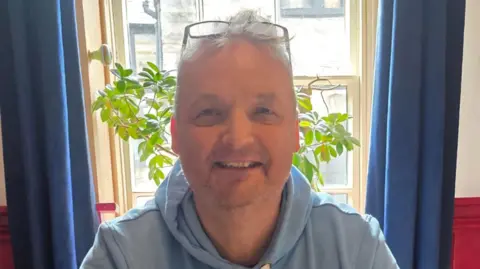 A man with short grey hair smiles towards the camera. He has a pair of glasses perched on top of his head and is clean shaven, wearing a blue hoodie. He is sat in front of a window with the sides of a plant poking into view either side of his head, with blue curtains either side.