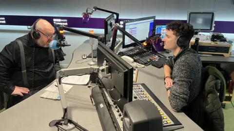 Bill Revans talks to Charlie Taylor in a radio studio. They are sat on opposite sides of a desk with microphones near their mouths.