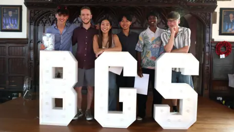 BGS Six A-level age students stand and smile in a group. In front of them are three large white letters - B, G and S