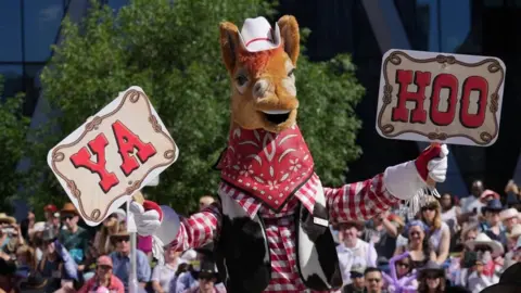Reuters Stampede mascot Harry the Horse displays signs during the Calgary Stampede parade. He wears a white cowboy hat with a thin red ribbon an da red plaid shirt with a cowhide sleeveless vest. He holds up two signs, one that reads 'YA' and the other that reads 'HOO'. 