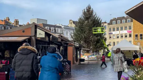 Visitors walking across the newly refurbished Northampton Market Square, featuring a large Christmas tree surrounded by fencing and a market stall displaying bags. 