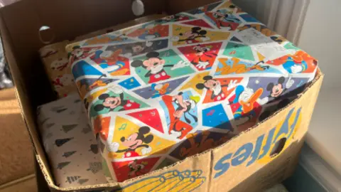 BBC A box of wrapped presents. The top parcel has Micky Mouse wrapping paper on it which is colourful. Below it is white wrapping paper with green Christmas trees on it. 
