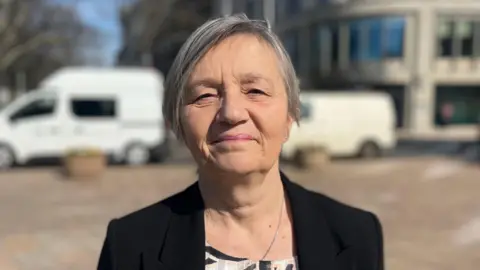 Jersey's Justice and Home Affairs Minister Mary Le Hegarat. She is stood on a street outside a building. Two white vans are behind her. The background is blurred. Le Hegarat has short grey hair and is wearing a black blazer with a white, grey and black dress underneath.