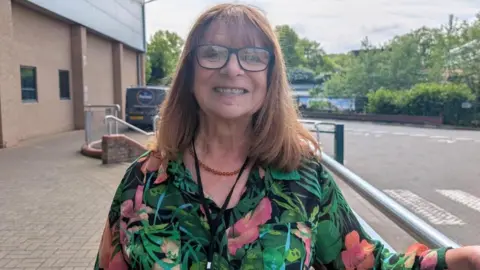 A woman with long brown hair, glasses and a green floral dress smiles as she leans on a hand rail outside a building.