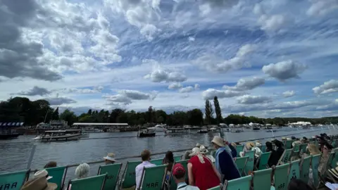 Sian & Poppy Cloudy skies hang above the River Thames at Henley as members of the regatta take to teal coloured deckchairs on the banks of the rover to watch the rowing races and small boats go past.