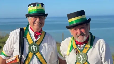 BBC/Will Flockton Two men wearing white shirts, black top hats decorated with green and yellow ribbons and similar colour ribbons around their chests smile into the camera.