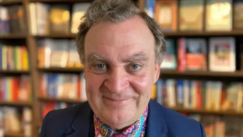 Author Dr John Llewellyn Probert smiling at the camera wearing a very colourful paisley shirt and tie with a dark blue suit